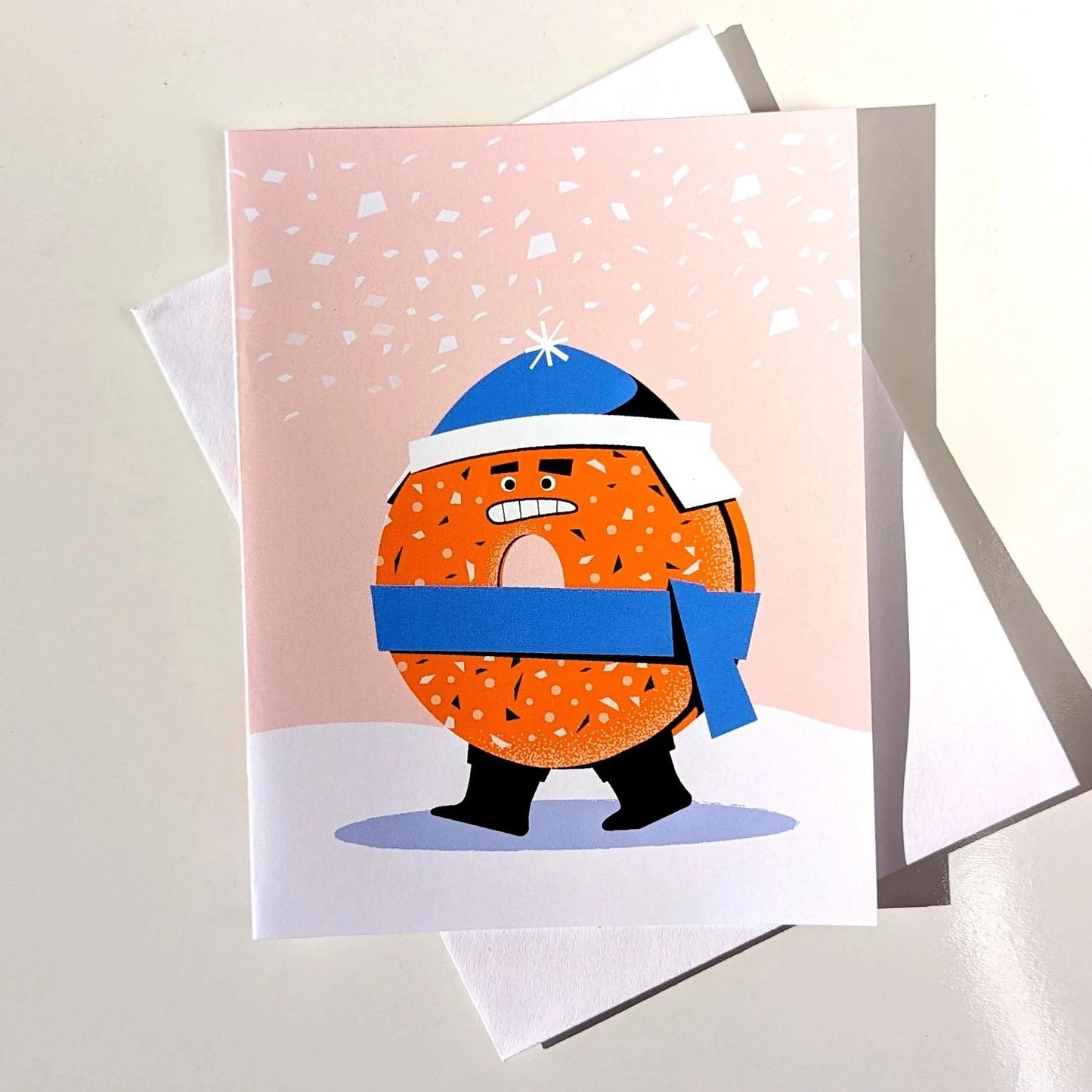 Greeting card with a cartoon character wearing a blue hat and scarf on a light pink background.